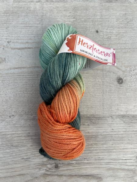 Garnstrang in Grüntönen und warmem Orange auf hellgrauem Holzuntergrund. Der Strang zeigt ein Farbverlaufspiel aus moosigen Grüntönen, petroligen Bereichen und einem kräftigen orangefarbenen Abschnitt. Die Banderole mit der Aufschrift „Herzfasern – Garnku